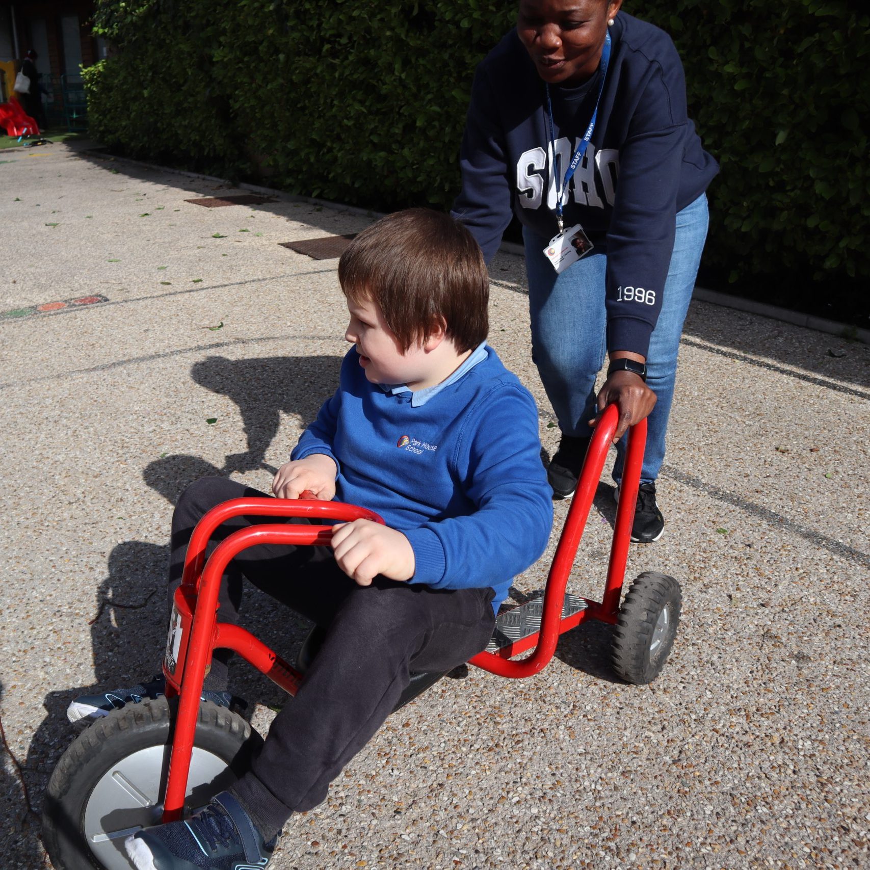 Boy riding red tricycle being pushed by woman